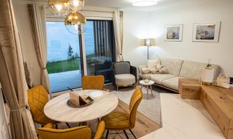 Modern living room with a comfortable couch, an elegant dining table, and yellow chairs. Large windows provide a view of a green garden outside.