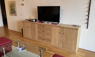 A modern living room with a wooden cabinet and a television. The floor is made of wood and there is a glass table with red chairs.