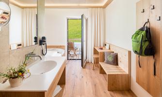 A modern bathroom with wooden and white elements. The door leads to a balcony with a view of nature.