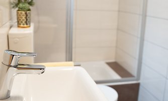 A modern bathroom with a shiny faucet and a white sink. In the background, a shower corner and a toilet room are visible.