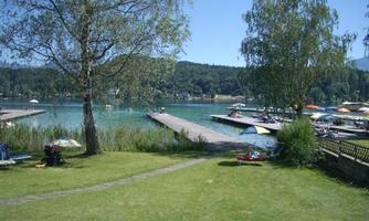 A tranquil lake with boat docks and green meadows. In the background, trees and sun umbrellas can be seen.