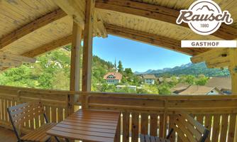 A cozy terrace with wooden furniture and a view of the surrounding mountains and the village. The landscape is green and inviting.