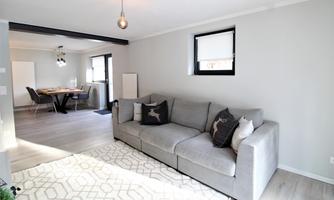 A modern living room with a gray sofa and decorative cushions. The room has light walls, an elegant carpet, and a dining area in the background.