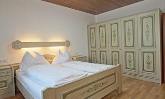 A cozy bedroom with a double bed and bright pillows. The room features a wooden floor and a large country-style wardrobe.