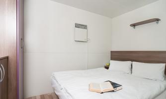 A simple bedroom with a double bed and a bedside table. An open book lies on the bed, and there is a small wall shelf.