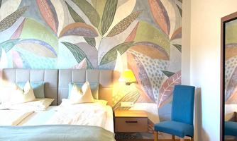 A cozy bedroom with a large bed and colorful wall design. There is a chair and a mirror that make the room inviting.
