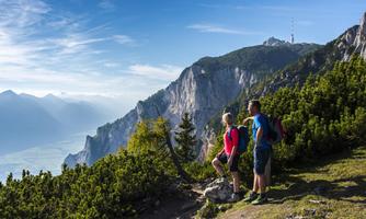 Two hikers enjoy the view of the mountains and the valley. The surroundings are green and sunny, with a clear blue sky.