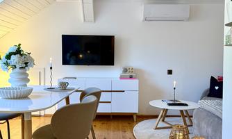 A modern living room with a dining table, cozy chairs, and a white wall. The room is bright and friendly, with a television and decorative elements.