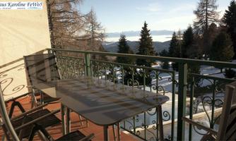 A balcony with a table and chairs, surrounded by a snowy landscape and mountains. In the background, trees are visible and the sky is clear.