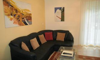 A modern living room with a black sofa and colorful cushions. Two pictures hang on the wall, and large windows let in plenty of light.