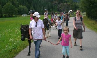 A group of children is riding on ponies and walking on a path. In the background, green meadows and trees can be seen.