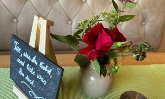 A table with a small board and a flower vase. The vase contains red flowers and is placed on a green cloth.