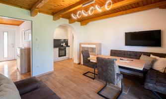A modern living room with a dining table and comfortable chairs. The open kitchen is well-equipped, and the space has a rustic charm.