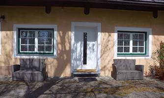 A cozy house facade with two pieces of seating in front of the door. The windows are framed in green and the walls are painted in a gentle shade of yellow.