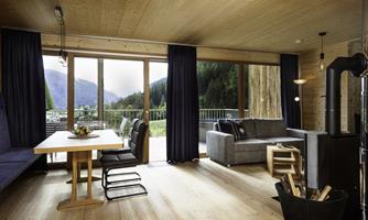 A modern living and dining room with wooden paneling and large windows. The room offers a view of the mountains and is cozy furnished.