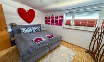 A cozy bedroom with a large bed and a red heart on the wall. The decoration is modern and inviting, with a carpet and windows that provide plenty of daylight.