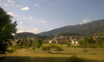 A picturesque landscape with gentle hills and a clear sky. In the foreground, meadows and trees are visible, and in the background, a small village.