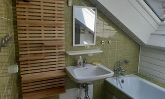 A modern bathroom with a sink, a bathtub, and a functional wooden shelf. The walls are tiled in a pleasant green tone, and a window provides daylight.