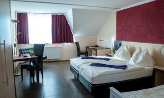 A bright room with two beds and a table. The windows are decorated with red curtains and the floor is tiled.