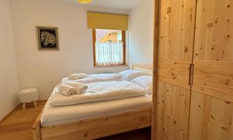 A cozy bedroom with a wooden double bed and soft colors.  
A window with a curtain lets in daylight and offers a view outside.