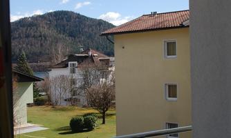 A view from a balcony overlooking a quiet residential area with green meadows and a hill in the background. The sky is blue and partly cloudy.
