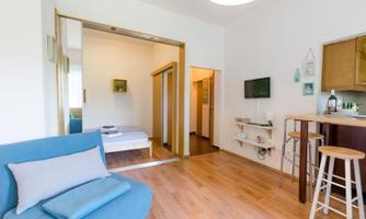 A bright and modern apartment with wooden flooring. The furnishings include a comfortable sofa, a small kitchenette, and a dining table.