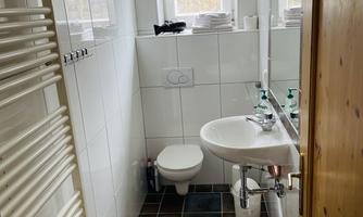 A small, modern bathroom with a sink, a toilet, and a window. The walls are tiled, and there are towels on a shelf.