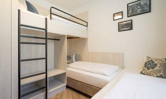 A modern bedroom with bunk beds and a single bed. The walls are decorated with pictures, and there is a simple wooden wardrobe.