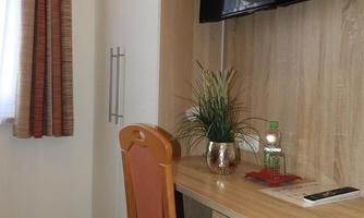 A simple desk with a chair is in a room, a television is mounted on the wall. Next to the desk is a plant and a few personal items.