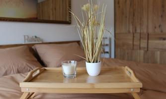 A wooden tray with a candle and a plant is placed on a bed with brown sheets. In the background, a wooden-paneled wall can be seen.