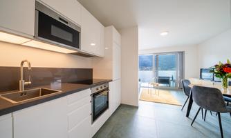 A modern kitchen with white cabinets and high-quality appliances. In the background, there is a dining area with a window and a view outside.