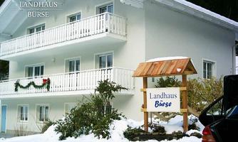 A cozy country house in the snow with several balconies and wooden signage. It stands surrounded by snowy ground and some plants.
