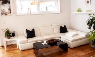 A modern living room with a white sofa and a black coffee table. The room has bright walls and a large window with natural light.