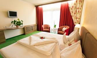 A cozy hotel room with a large bed and fresh flowers on the table. The windows are decorated with red curtains and let in plenty of daylight.