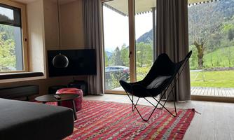 A modern, cozy living room with large windows and a view of the green landscape. The room is furnished with a colorful rug, a chair, and a television.