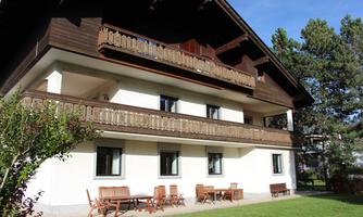 A modern house with two floors and several balconies. The garden is well-kept and features wooden seating furniture.