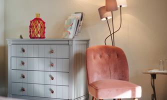 A modern room with a gray dresser and a pink armchair. Next to the armchair stands a floor lamp with a warm light.
