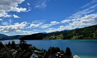 A picturesque landscape with a clear lake and gentle hills in the background. The sky is bright blue and adorned with white clouds.