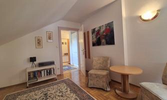 A cozy room in the attic with two chairs and a small table. A painting hangs on the wall and there is a bookshelf as well as a rug on the floor.