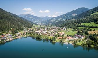 A picturesque village on the shore of a clear lake, surrounded by green mountains. The sky is blue with some clouds.