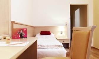 A simple room with a single bed and a desk. There is a chair and a small bedside table with a lamp.