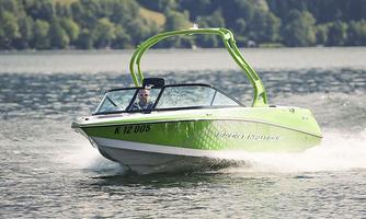 A green motorboat is speeding across a clear lake. In the background, green trees and hills can be seen.