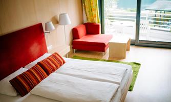 A modern bedroom with a red headboard and striped pillows. In the room, there is a red sofa and there are large windows with a view outside.