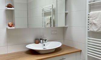 A modern bathroom with a white wall and a wooden vanity. In the background, a large mirror reflects, and a towel warmer is visible.