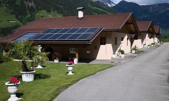 A modern house with solar panels in a mountainous landscape. The path is lined with greenery and flower beds.