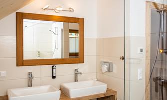 A modern bathroom with two sinks and a large mirror. The walls are light-colored and the shower area is separated by a glass wall.