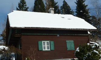 A traditional wooden house in the snow with green shutters. In the background, there are tall fir trees and a clear sky.