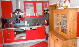 A modern kitchen with red furniture and stainless steel appliances. A wooden cabinet with glasses stands in the corner.