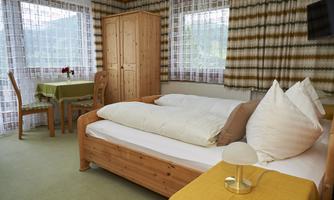 A cozy room with a wooden bed, a table, and chairs. Large windows let in a lot of light, and the decoration is simple and friendly.