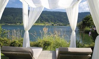 A relaxing view of a lake with gentle hills in the background. Two comfortable loungers are positioned under a white canopy.